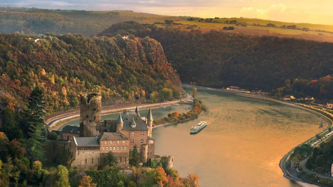 Katz Castle overlooks the winding Rhine River in Germany's Middle Rhine Valley, surrounded by forested hills in autumn colours as a river ship cruises below at sunset.