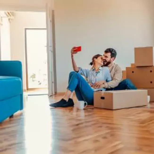 Hew homeowner couple sits on the floor of their living room o take a selfie between boxes