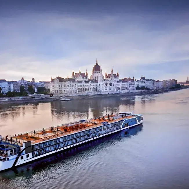 River cruise ship sailing on a calm river with a grand historic building along the shoreline.