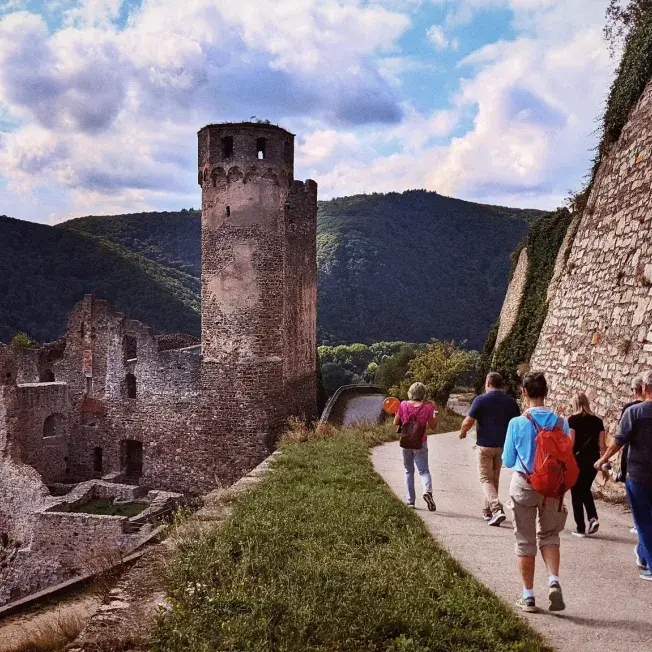 People walking on a riverside path beside castle ruins and stone walls.