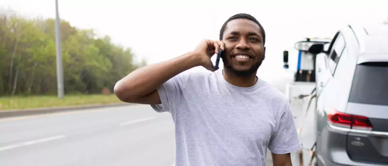 Man smiling with a phone to his ear with a car on a tow truck in the background