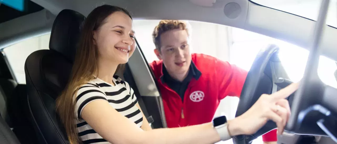 Young female in a stripe tshirt in the driver seat of a car touching the navigation screen with a CAA worker looking through her window asissting