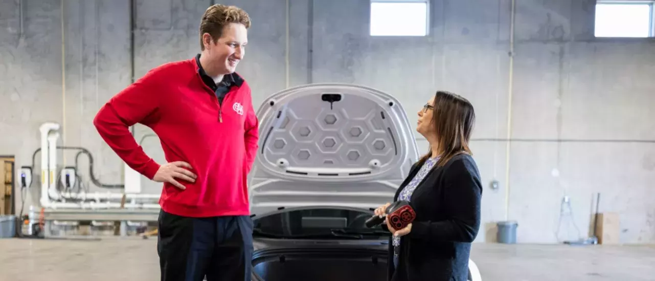 A man in a red CAA sweater looking at a woman holding an electric vehicle charger, both standing in front of a car with its hood up