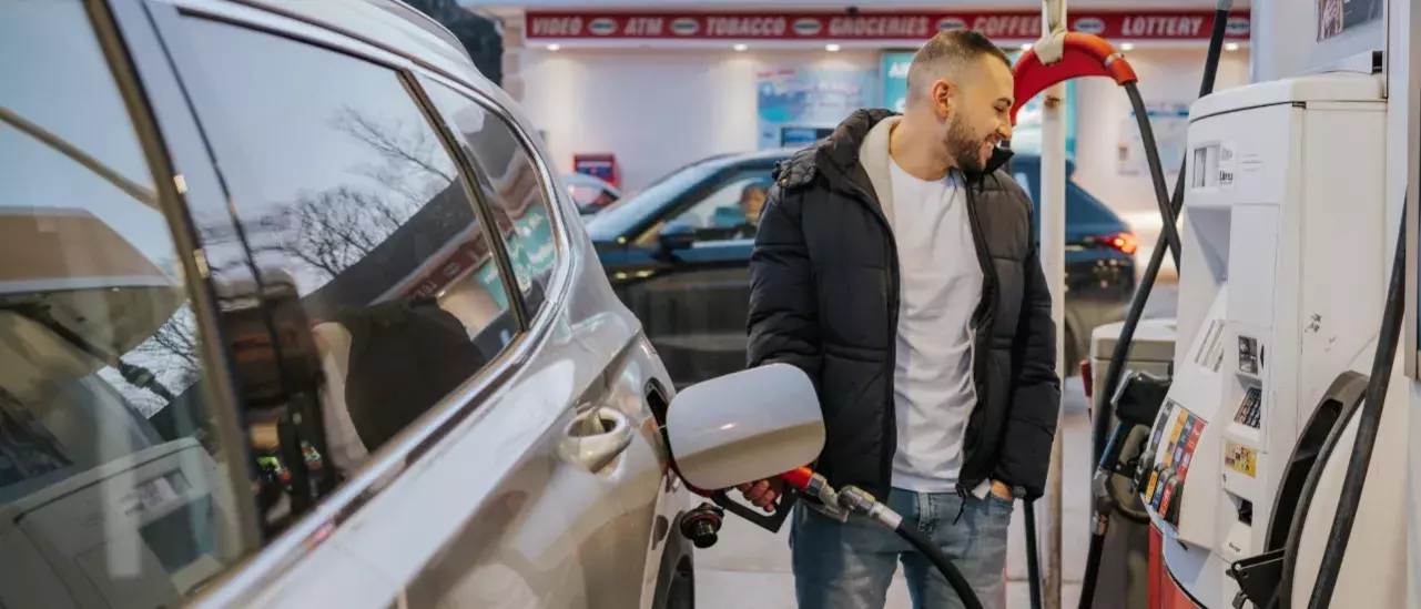 Man in black winter jacket, white tshirt and jeans fueling his silver SUV at a gas station smiling looking at the pump 