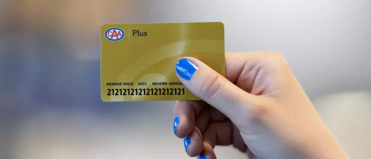 hand with blue nailpolish holding a gold CAA membership card with the word Plus at the top left to the right of the CAA logo