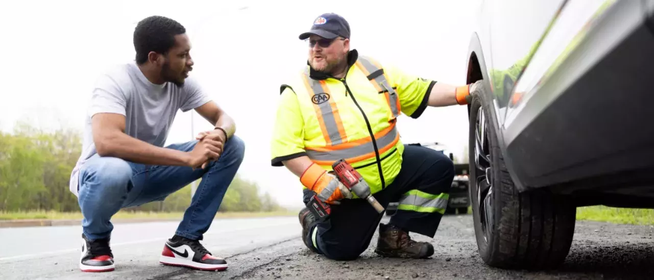 Two males crouching beside a vehicle, one being a CAA roadside attendent holding a power tool by a tire