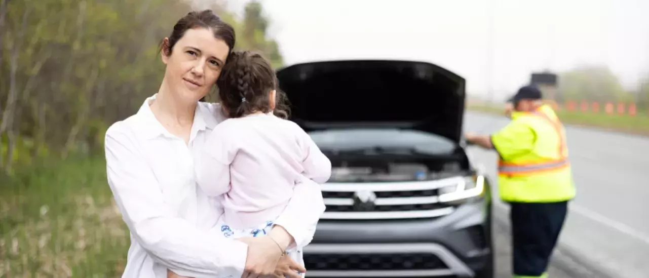 Young mother wearing a white button down shirt holding a small child who is looking back at their vehicle that has the hood open that is being serviced by a roadside technician in neon reflective gear