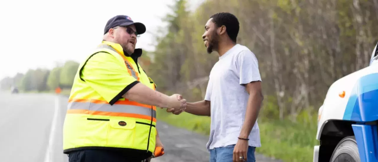 Roadside assistance worker in a high-visibility jacket shakes hands with a man beside a CAA-branded truck on the roadside.