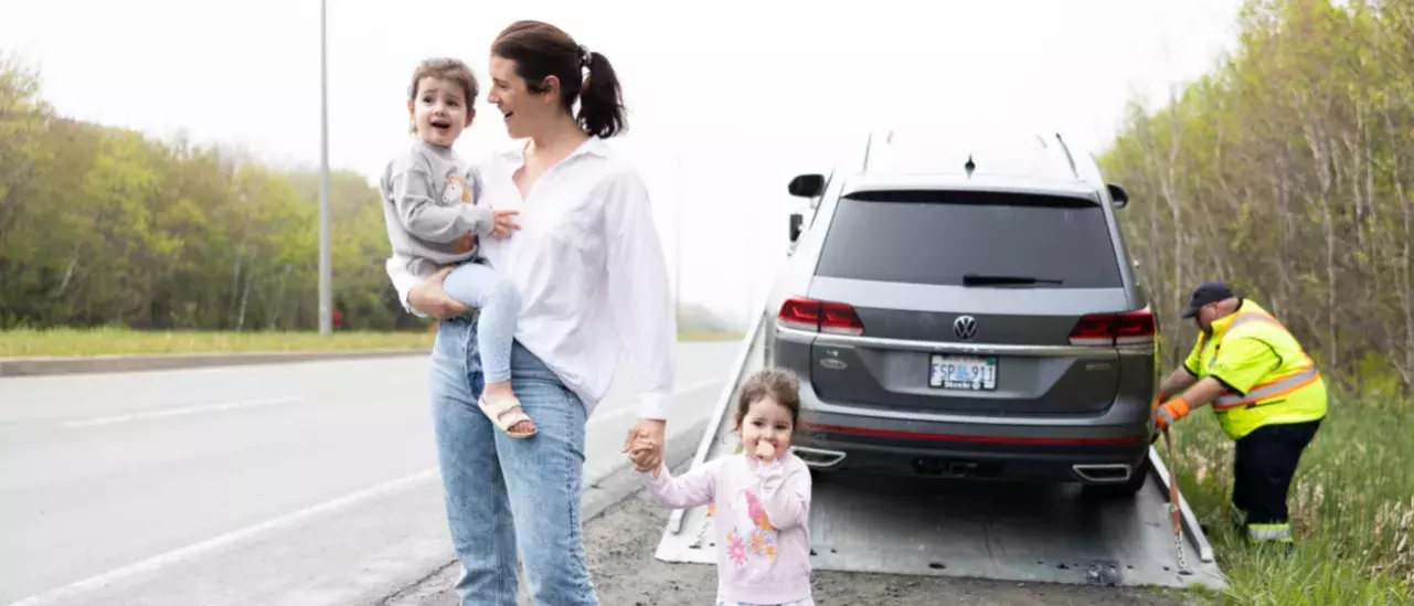 young caucasian mother holding one child in her arms and holding another childs hand on the shoulder of a highway. Pictures behind her is a Volkswagen SUV being secured on a tow truck by a roadside assistance worker in neon saefty gear