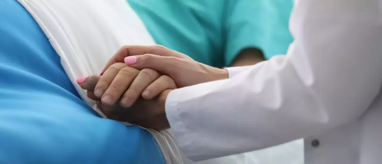 close up shot of a doctors hand with pink nailpolish holding a patients hand while they are laying in a bed with a blue blanket