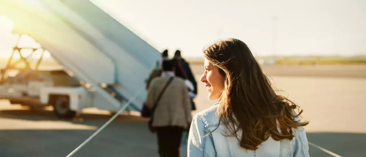 Woman wearing a jean jacket smiling and looking to her left as she approaches a plane wiht passengers heading towards the stairs