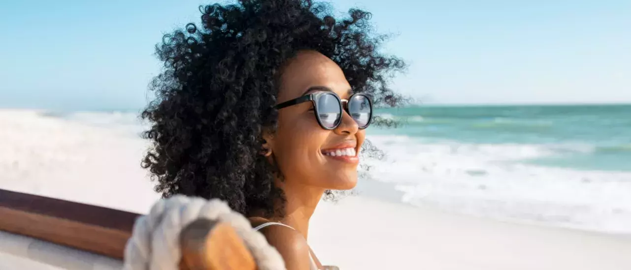 close up shot of a woman smiling with curly hair wearing black sunglasses sitting on a beach chair at the beach 