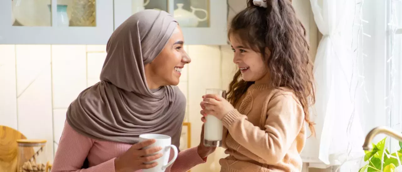 Young mother wearing a pink long sleeve shirt and a grey hijab holding a white coffee cup smiling at a young girl wearing a camel sweater holding a glass of milk in their kitchen