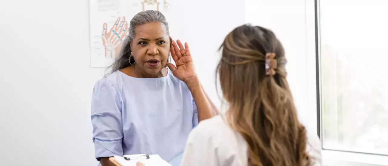 Lady in a doctors office in a blue dressing gown holding up a hand to her ear to a doctor with a clipboard