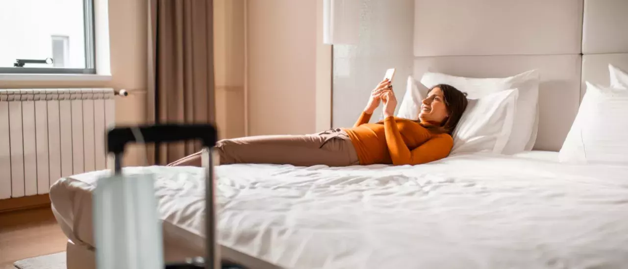 Woman in a hotel room wearing an orange turtleneck and khaki pants laying on the bed smiling at her phone