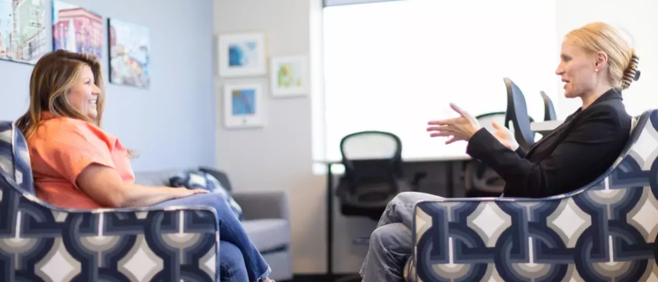 Two females sitting in arm chairs facing each other, smiling, having a conversation