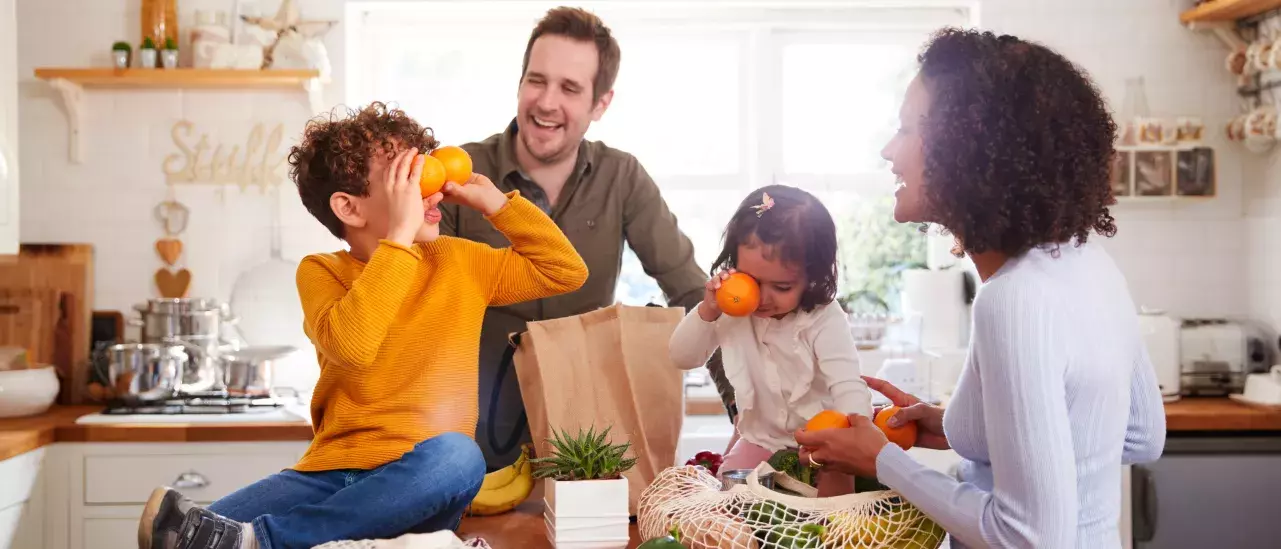 A family of four is in the kitchen, unpacking groceries. Two children play with oranges, while a man watches and a woman smiles. The scene is bright, with groceries and a plant on the counter.