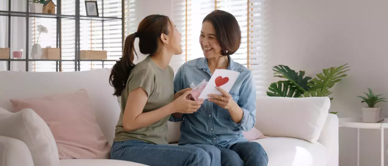 Two people sitting on a couch, smiling and exchanging greeting cards. One person is holding a card with a red heart on it. Room with light decor and plants in the background.