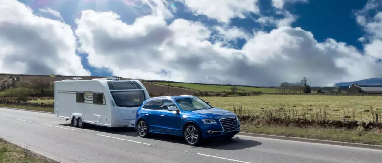 Blue SUV is towing a family camper down a highway surrounded by grassy fields on a sunny day. 