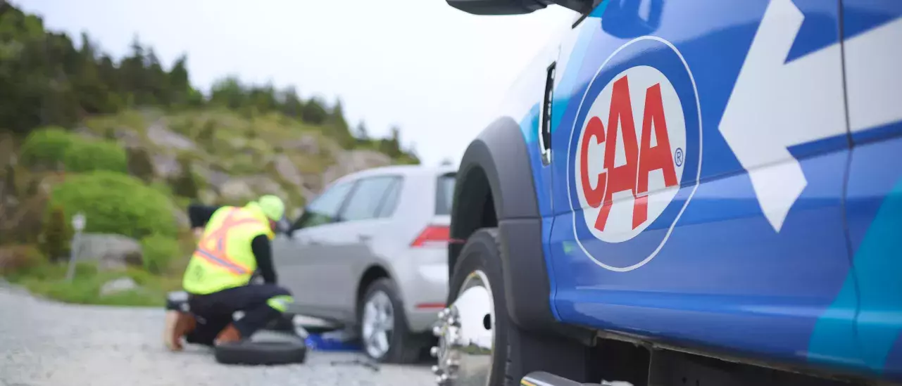 A CAA roadside service vehicle is parked next to a car receiving assistance. The technician, wearing a safety vest, is inspecting the car's front tire with tools on the ground.