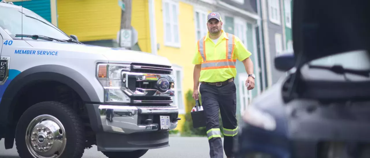 A roadside assistance worker in a high-visibility yellow shirt and cap walks toward a vehicle with an open hood. A service truck with "Member Service" written on the door is parked nearby. Colorful houses line the street in the background.
