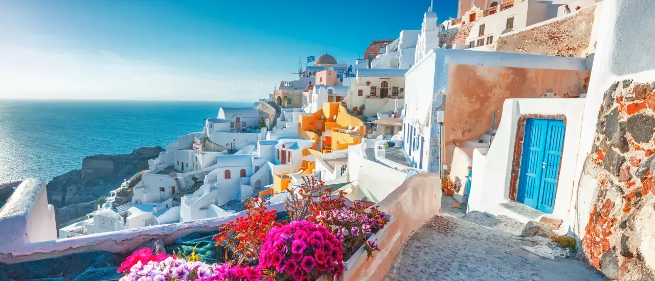 A picturesque view of Santorini, Greece, featuring white-washed buildings with blue domes cascading down a hillside. Vibrant pink flowers in the foreground contrast with the deep blue sea and sky, highlighting the island's charming architecture and stunning scenery.