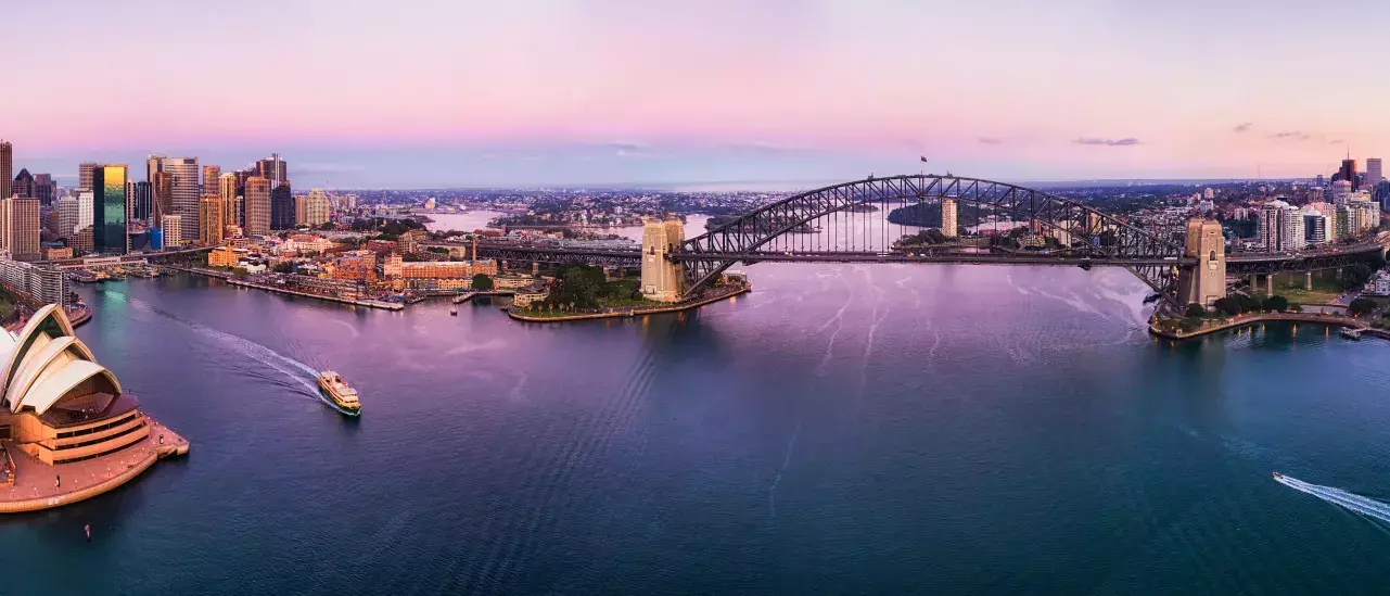 A panoramic aerial view of Sydney Harbour at sunset, featuring the Sydney Opera House on the left and the Sydney Harbour Bridge in the center. The city skyline is visible with numerous buildings, and the sky is painted with pink and purple hues.