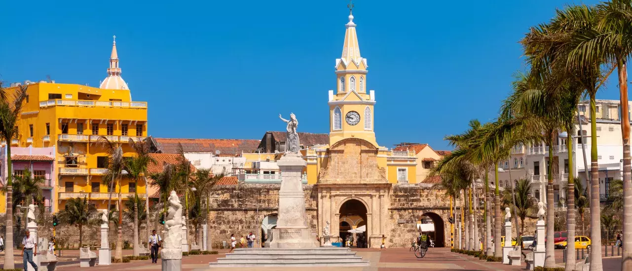 A city plaza featuring a clock tower, several statues, palm trees, and surrounding buildings under a clear blue sky.