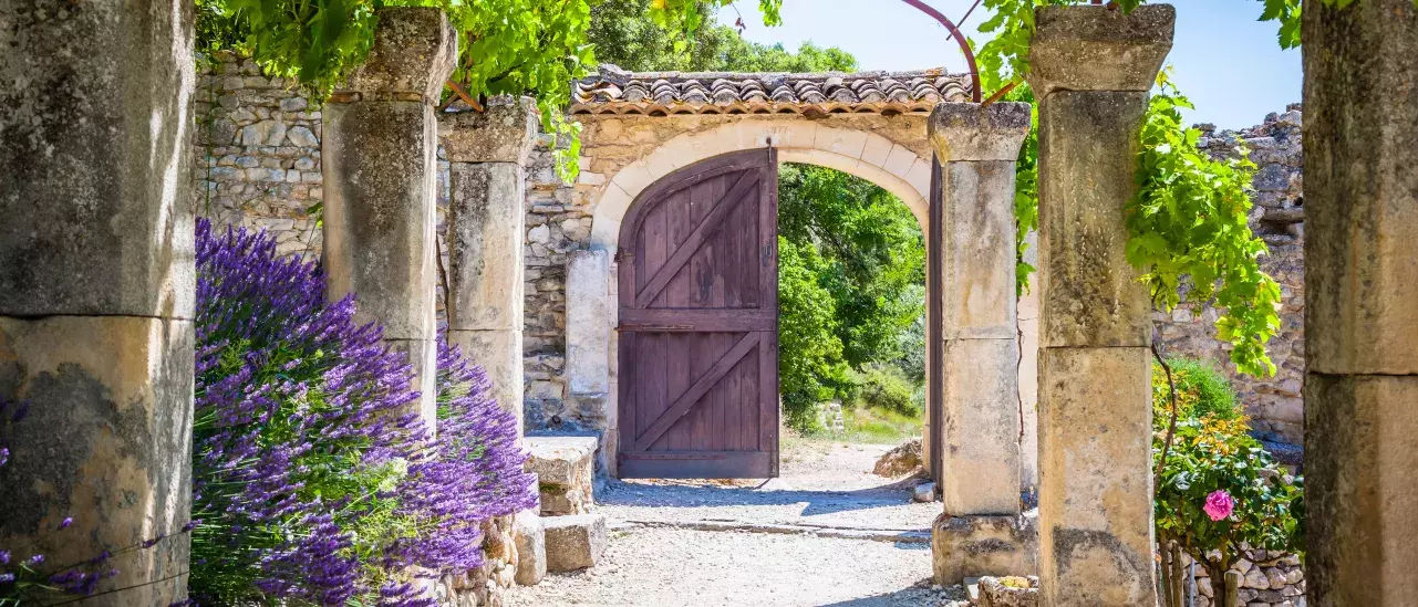 A rustic stone archway with a wooden door, surrounded by lush green vines, lavender bushes, and stone pillars, leading to an outdoor area on a sunny day.