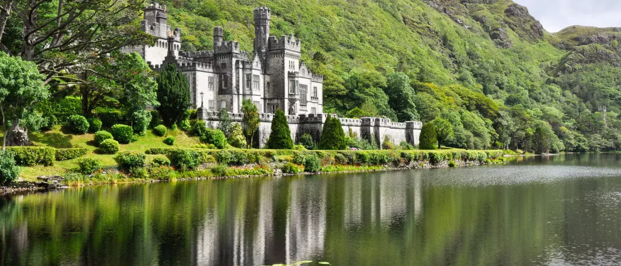 A castle with towers and turrets is situated beside a lake, surrounded by lush greenery and hills.