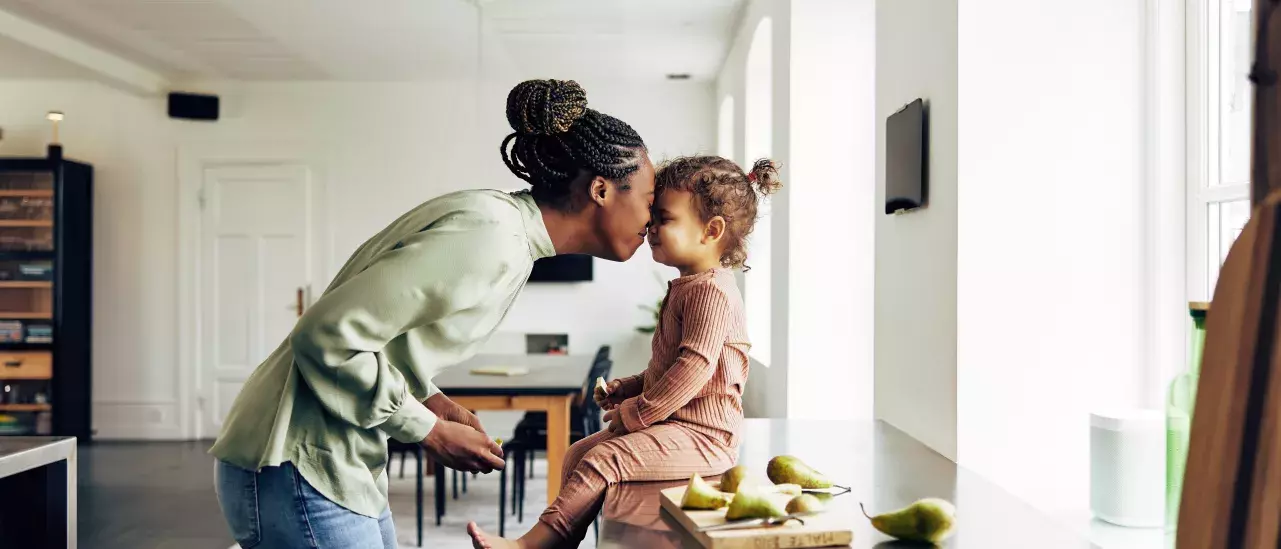 A mother gently kisses her young child on the forehead as the child sits on a kitchen counter. 