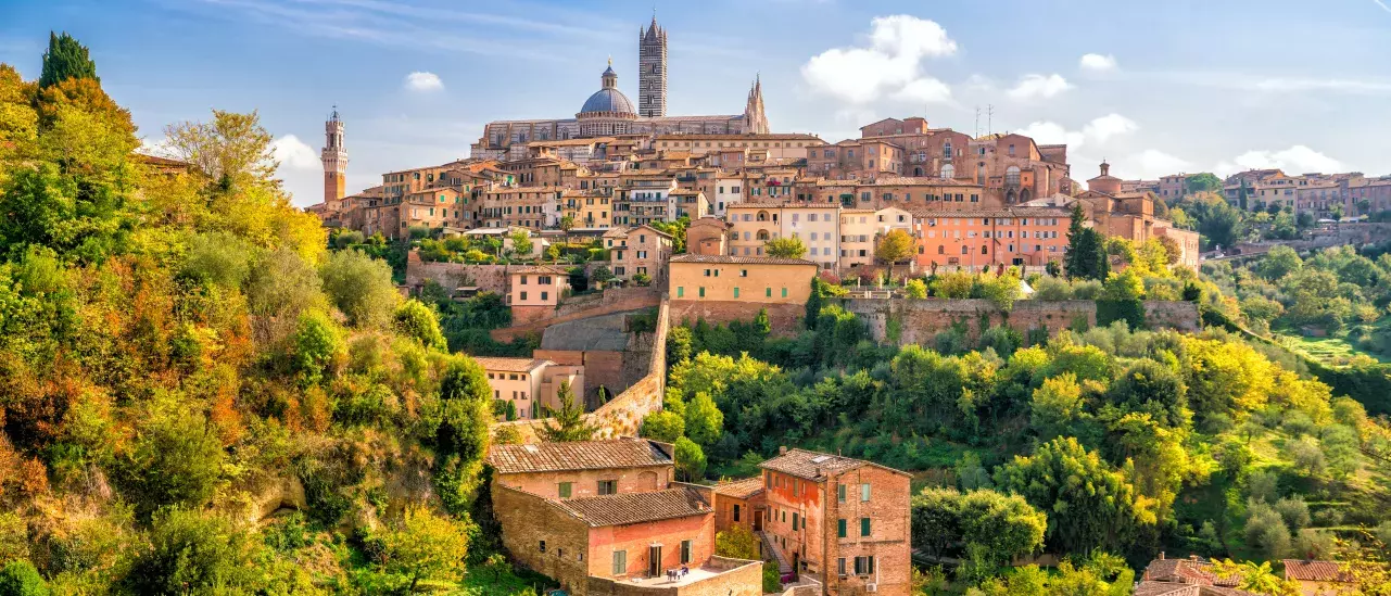 A scenic view of Siena, Italy, featuring a medieval hilltop town with terracotta-roofed buildings, lush greenery, and a clear blue sky. 