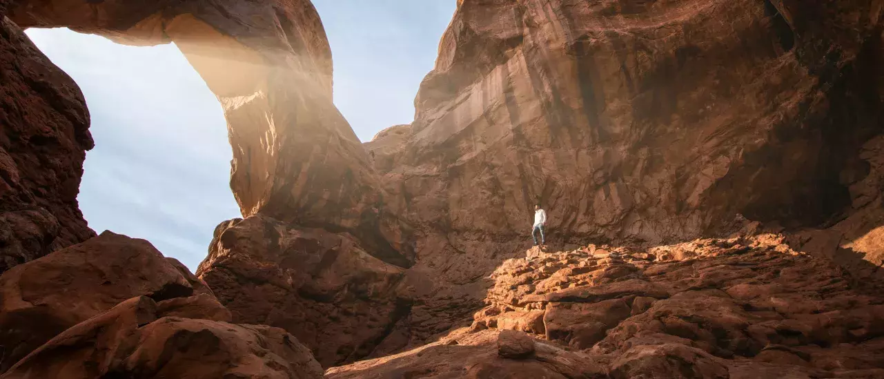 A person stands alone on a rocky terrain, dwarfed by a massive natural rock arch illuminated by soft sunlight streaming through the gap.