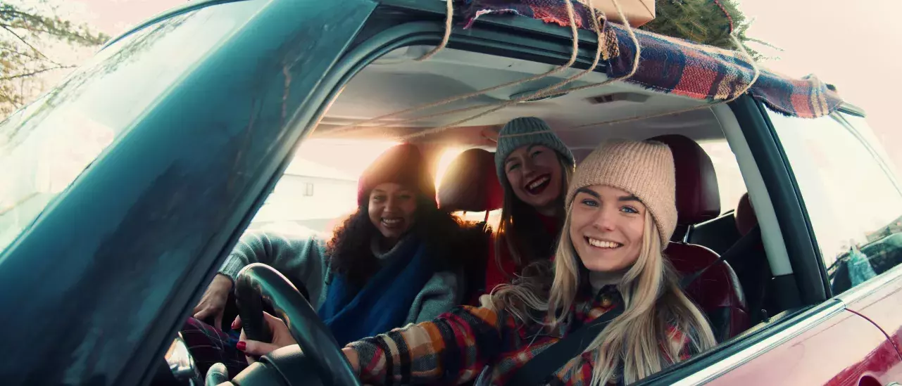 Three friends wearing winter hats, smiling in a car packed with holiday items and a Christmas tree on the roof.