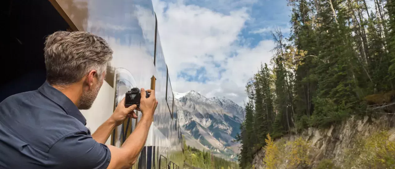 A man leaning outside of the Rocky Mountaineer train with a camera taking a picture of the Rocky Mountains.