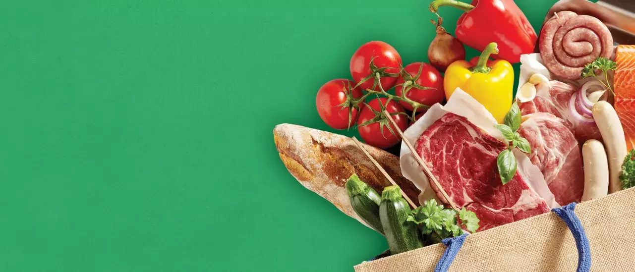 A grocery bag filled with various fresh food items, including tomatoes, a yellow bell pepper, a loaf of bread, zucchini, leafy greens, raw meat cuts, sausages, and cheese. 