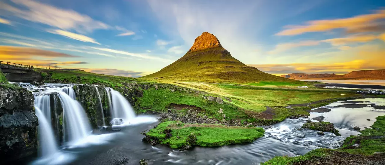 A cone-shaped mountain with a grassy foreground and a waterfall cascading into a river on the left. The sky is clear with some clouds illuminated by the setting or rising sun.