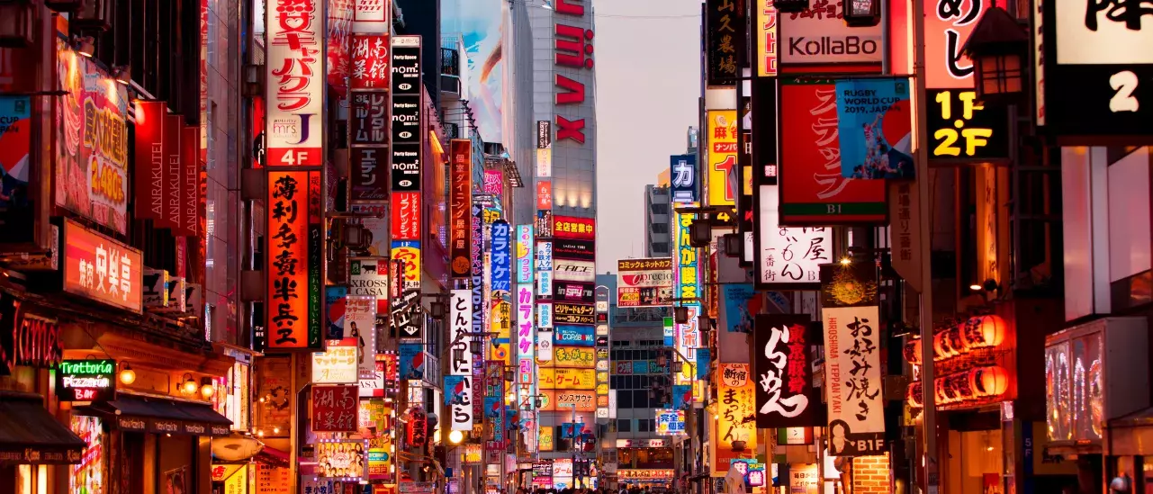 A bustling street in Japan filled with numerous brightly lit signs and advertisements in Japanese. The street is crowded with people walking, and the buildings are adorned with colorful neon lights and billboards.