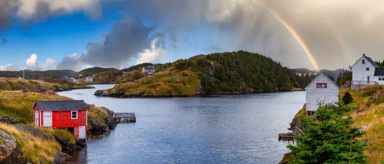 A picturesque coastal scene with a small inlet of water surrounded by grassy hills and scattered houses. A red wooden house is near the water's edge on the left, while white wooden houses are on the right. A rainbow arches across the sky above the hills in the background, under a partly cloudy sky.