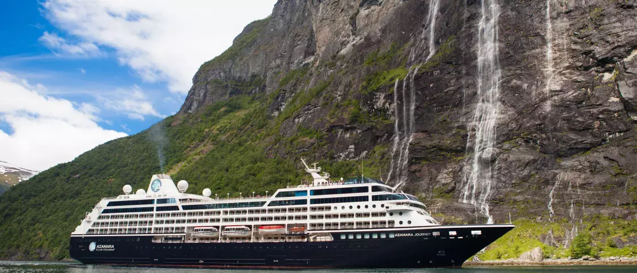 A large cruise ship sailing near a steep, rocky cliff with multiple waterfalls cascading down the cliffside. The water is calm, and the sky is partly cloudy with patches of blue. The ship has several decks and various structures on top, including domes and antennas.