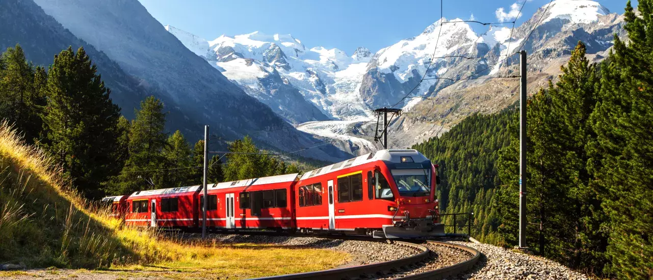 A red train traveling on a curved railway track through a mountainous landscape with snow-capped peaks, green trees, and a clear blue sky with scattered clouds.