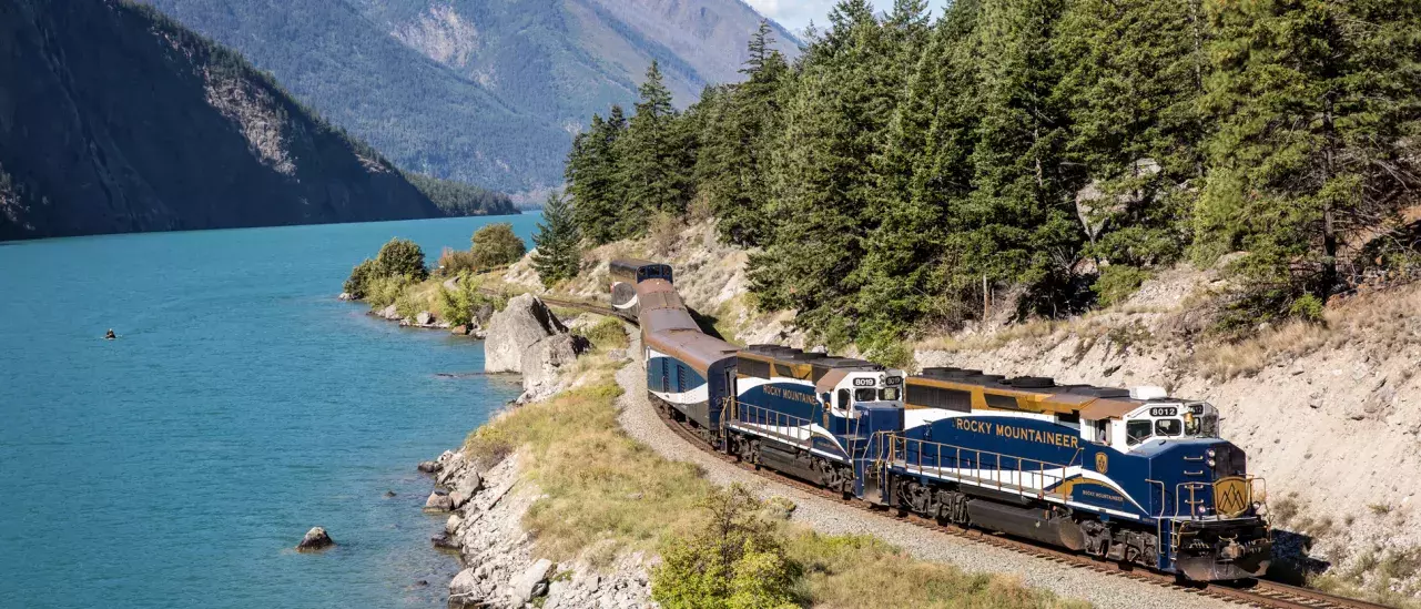 The Rocky Mountaineer train traveling along a scenic route beside a turquoise lake, surrounded by lush green trees and mountains in the background. The train is blue with white and gold accents, and the words "ROCKY MOUNTAINEER" are visible on the side of the lead locomotive.