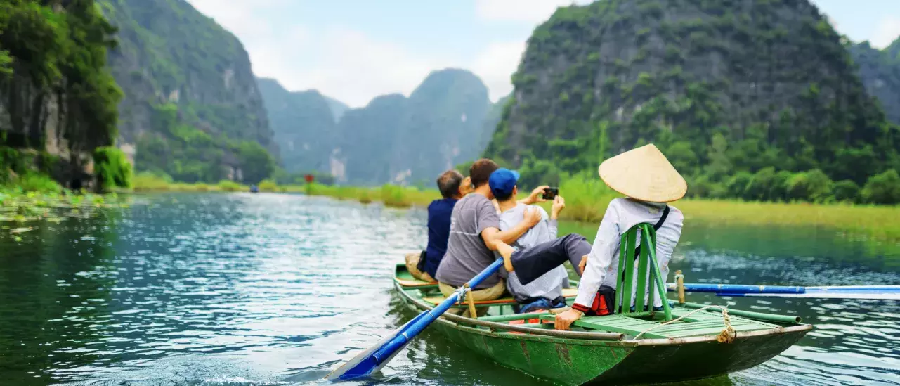 Four people in a green boat on a calm river, surrounded by lush green mountains. One rows with blue oars, another wears a conical hat, and two take photos.