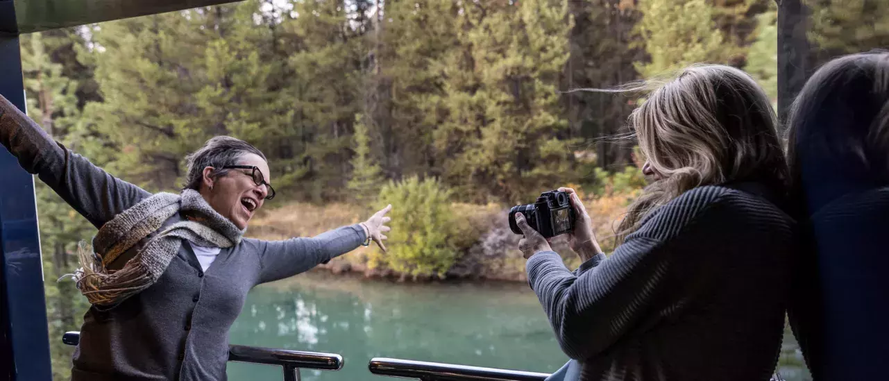 Two people on a balcony overlooking a river and forest; one poses while the other takes a photo.