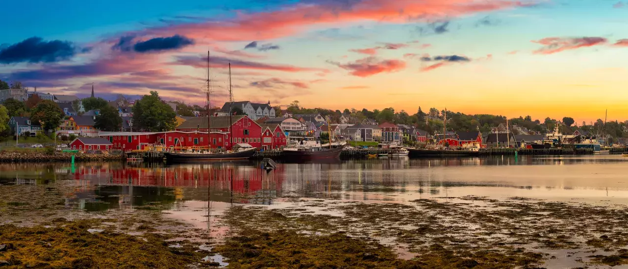 Sunset over a coastal town with colorful buildings, boats docked along the calm waterfront, and a rocky shoreline in the foreground.