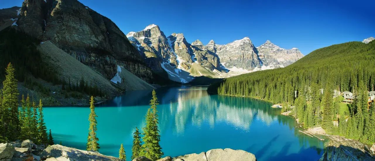 A serene turquoise lake surrounded by dense evergreen forests and towering snow-capped mountains under a clear blue sky.