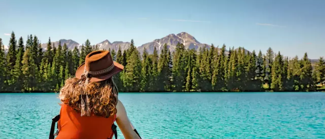 A person with curly hair, wearing a brown hat and an orange sleeveless top, is paddling a canoe on a turquoise lake. The person is facing away from the camera towards a dense forest of evergreen trees and distant mountains under a clear blue sky.