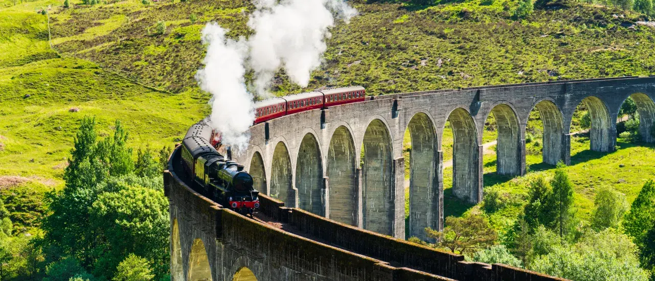 Steam train crossing a curved viaduct in a green hilly landscape.
