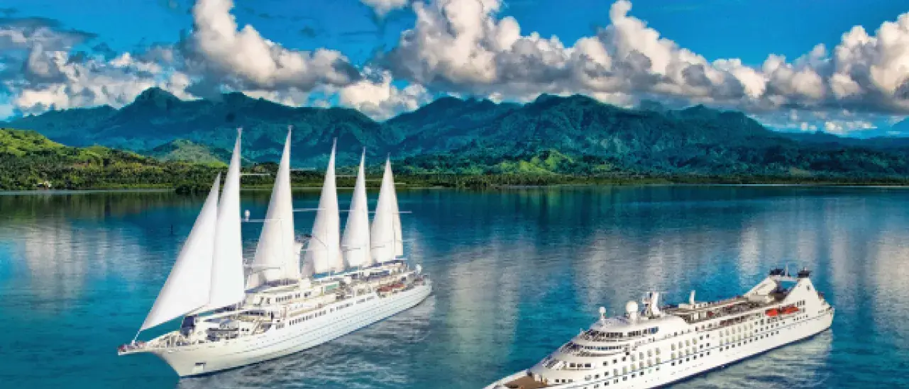 Two large cruise ships sail on calm, clear water surrounded by lush green mountains and a partly cloudy sky. Tropical palm trees frame the foreground. One ship features multiple sails, while the other is a modern cruise liner.