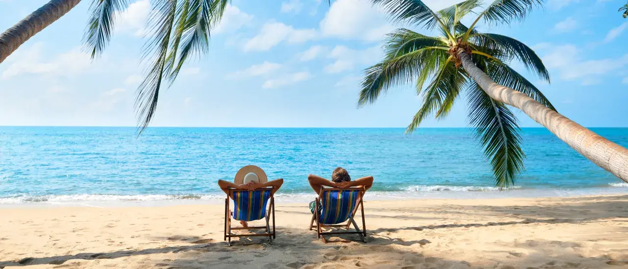 Two people lounge on beach chairs under palm trees by the sea, enjoying a sunny tropical day.